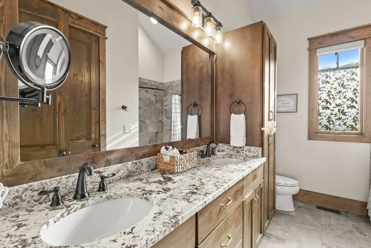 A luxurious bathroom with double sinks set in a granite countertop, wooden cabinetry, decorative lighting fixtures, and a window with patterned glass. The ambiance is calm and rejuvenating, ideal for self-care routines.