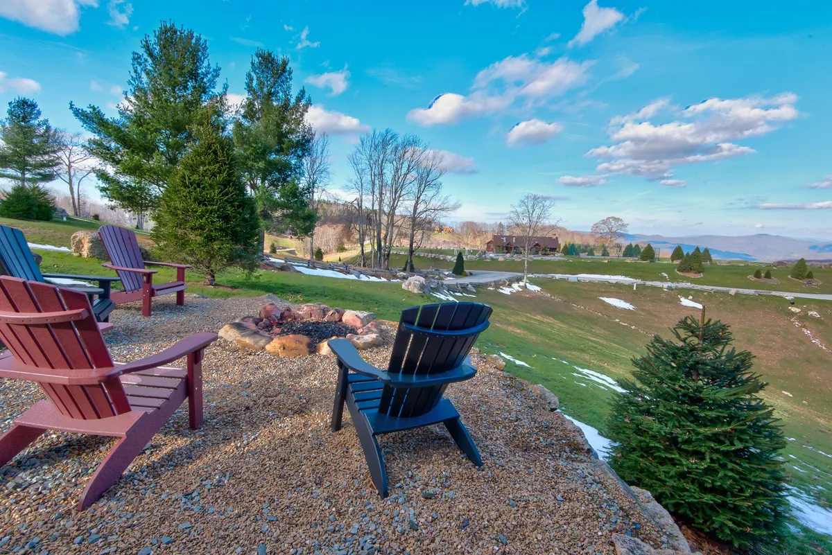 A grouping of Adirondack chairs encircles a fire pit on a lush, green lawn with distant mountains in the background. The scene is relaxed and social, ideal for stargazing and sharing stories by the fire with friends or family.
