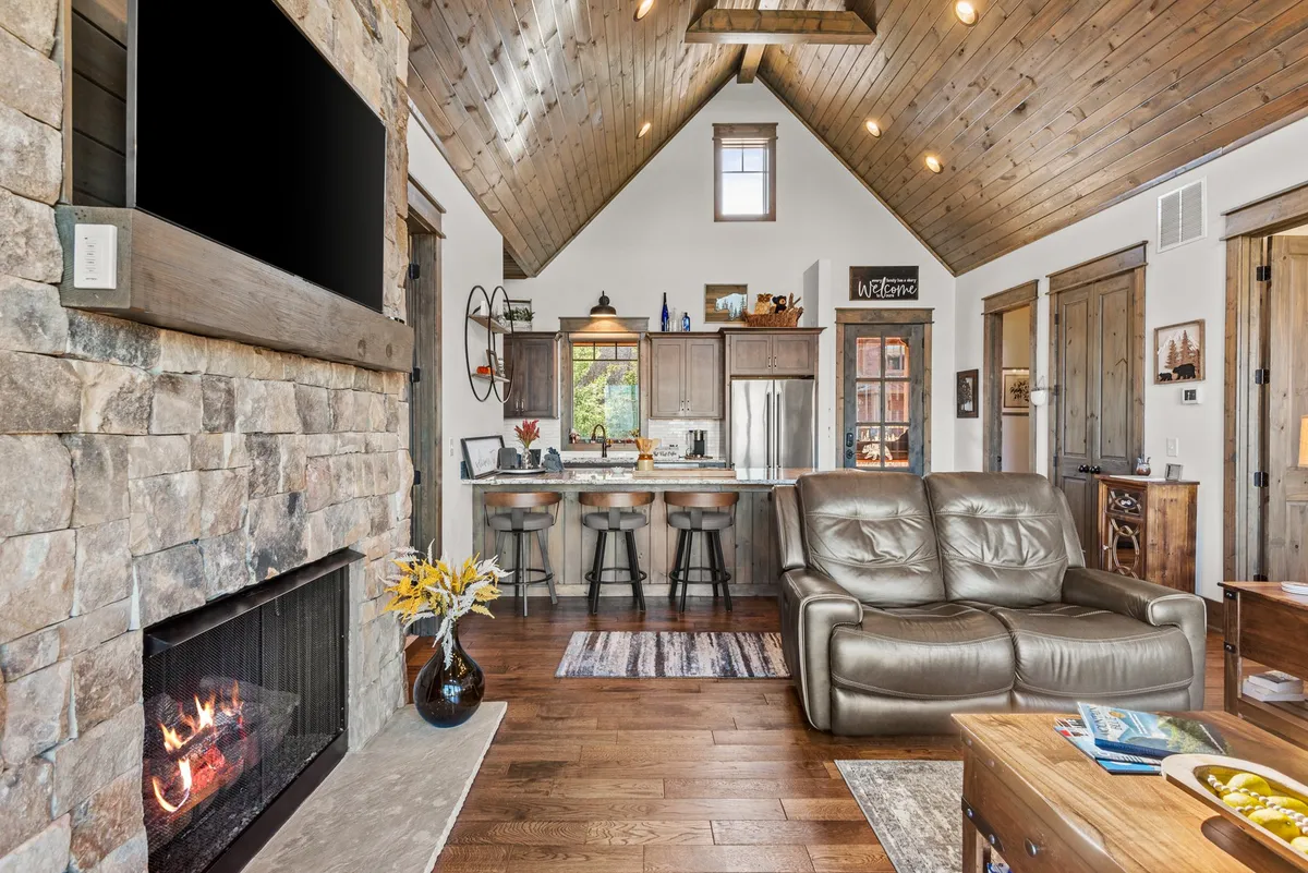 The image features a spacious, open-plan area with a comfortable leather sofa, kitchen island with bar stools, stone fireplace, and large windows bathing the room in natural light. The kitchen and dining zones flow seamlessly together, promoting social interaction.