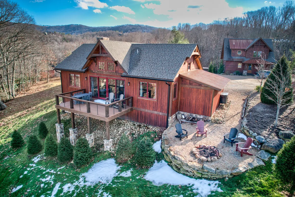 The image showcases a charming wooden cabin nestled in the natural landscape of Banner Elk, North Carolina. The inviting deck is set up with comfortable seating, overlooking breathtaking sunset views. A cozy fire pit area beckons guests to gather outdoors, while the peaceful surroundings highlight the opportunity for nature walks and tranquil evenings under the stars.
