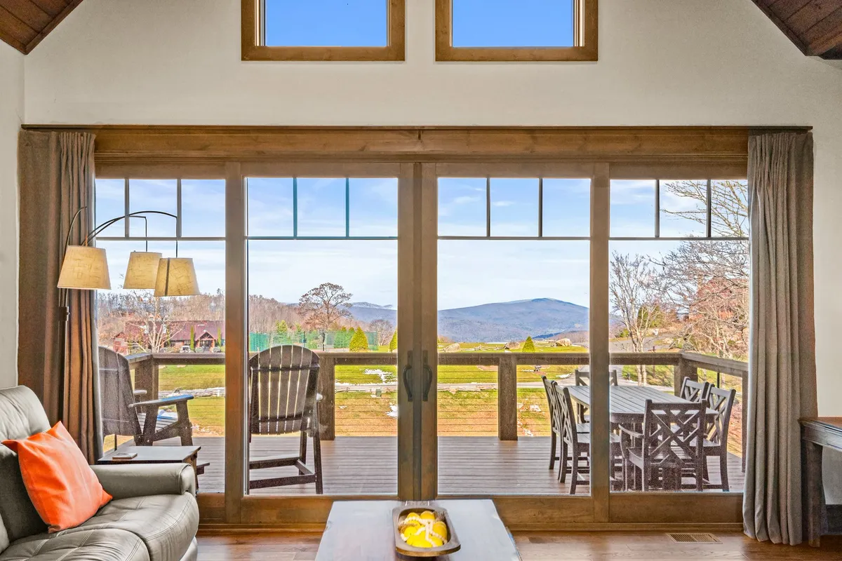 A cozy interior scene with large sliding glass doors opening onto an outdoor deck, comfortable seating, and a bright, inviting atmosphere. The design draws attention to indoor-outdoor living, with views of the natural landscape beyond the windows.