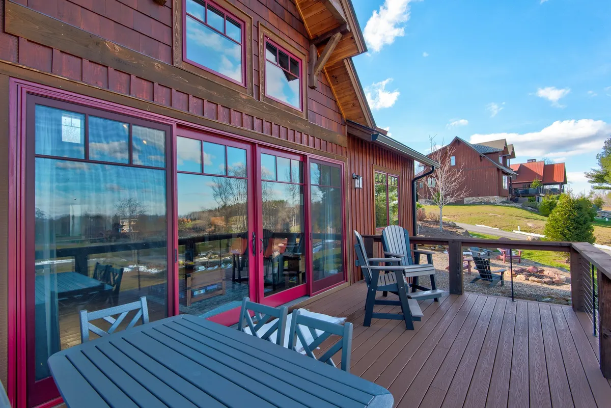 This image shows a welcoming exterior view of Sommer Hus, featuring large windows that allow plenty of natural light inside, inviting deck seating, and a landscaped yard. The deck is easily accessible and offers generous space for relaxing or entertaining while enjoying beautiful sunset views and morning coffee overlooking the landscape.