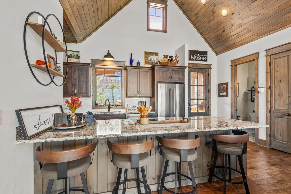 This image displays a kitchen island with a granite countertop, surrounded by bar stools. The kitchen features wooden cabinetry, decorative shelves with items, and a 'welcome' sign, all warmly illuminated. The scene conveys a sociable, inviting environment ideal for both cooking and gathering.