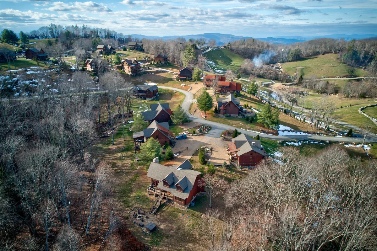 A row of rustic cabins set against a mountain backdrop, with winding roads, lush greenery, and scattered snow patches. The scene speaks to outdoor exploration, family retreats, and the changing seasons, inviting guests into a peaceful communal environment.