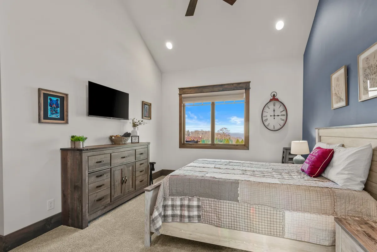 A serene bedroom featuring a bed with a patchwork quilt, a large window showcasing scenic views, a wooden dresser adorned with decorative items, and a wall-mounted Smart TV.