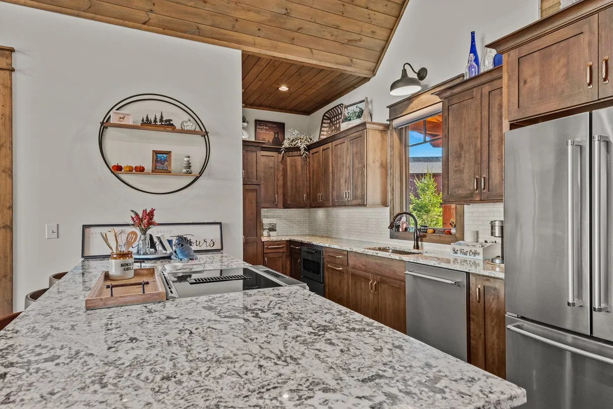 A cozy kitchen with a prominent granite countertop island, wooden cabinets, and a window letting in natural light. The decorative shelf with curated items adds a personal touch. The layout encourages interaction and togetherness, ideal for family gatherings or meal prep.