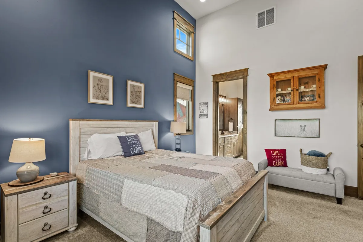 A calm and comfortable bedroom with a bed dressed in a patchwork quilt, decorative pillows, a nightstand with a lamp, framed artwork, and windows letting in natural light. The room features a doorway that leads directly to an en-suite bathroom.