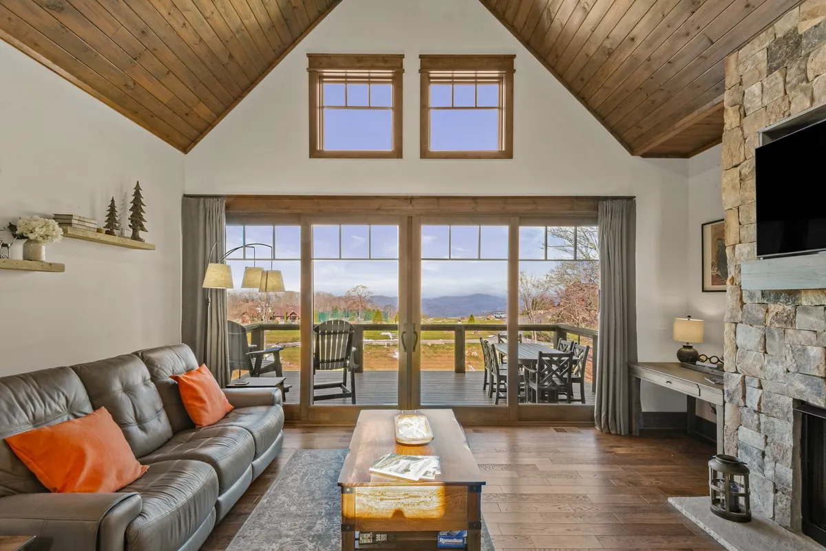 This image captures a cozy living room featuring a plush leather sofa arranged near a striking stone fireplace. Large windows dominate one wall, flooding the space with natural light and offering panoramic mountain views. A coffee table sits in the center, perfect for gatherings, and the overall ambiance is warm and inviting, accentuated by the wood floors and high ceiling.