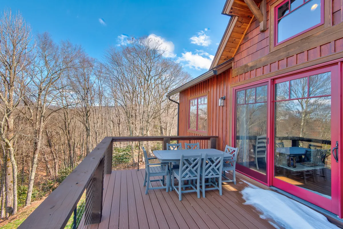 A spacious wooden deck with an outdoor dining set is surrounded by tall trees and features large glass windows leading into the home. The deck is set up for relaxing and gathering with friends in an inviting atmosphere, with nature all around and glimpses of the sky above.