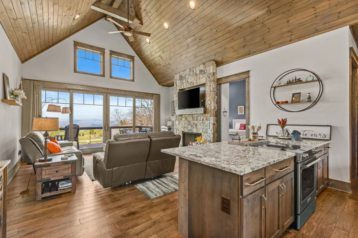 A spacious, open-concept living area with a modern stone fireplace, kitchen island with granite counter, and large windows showcasing the scenic outdoors. The hardwood floors and cathedral ceiling contribute to the open, airy feel, while the sightlines between kitchen and living space foster togetherness.